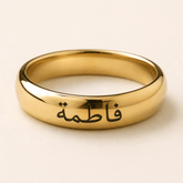 A polished gold ring engraved with the Arabic name 'Fatima' in black script, shown on a light beige background.
