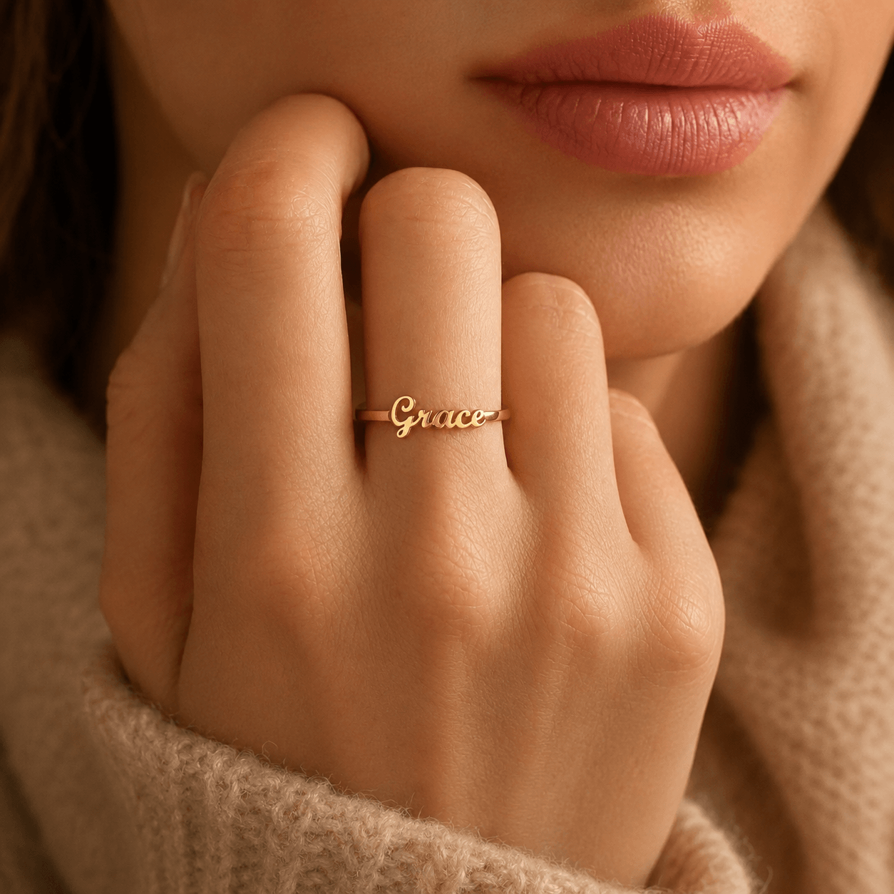 A close-up lifestyle shot of a woman's hand resting near her chin, wearing a delicate gold script ring that reads 'Grace'. She is wearing a soft beige knit sweater, and the background is a warm, softly blurred neutral tone. from shine gold and diamonds