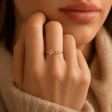 A close-up lifestyle shot of a woman's hand resting near her chin, wearing a delicate gold script ring that reads 'Grace'. She is wearing a soft beige knit sweater, and the background is a warm, softly blurred neutral tone. from shine gold and diamonds