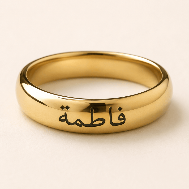 A polished gold ring engraved with the Arabic name 'Fatima' in black script, shown on a light beige background.