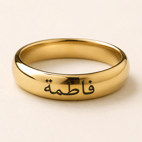 A polished gold ring engraved with the Arabic name 'Fatima' in black script, shown on a light beige background.