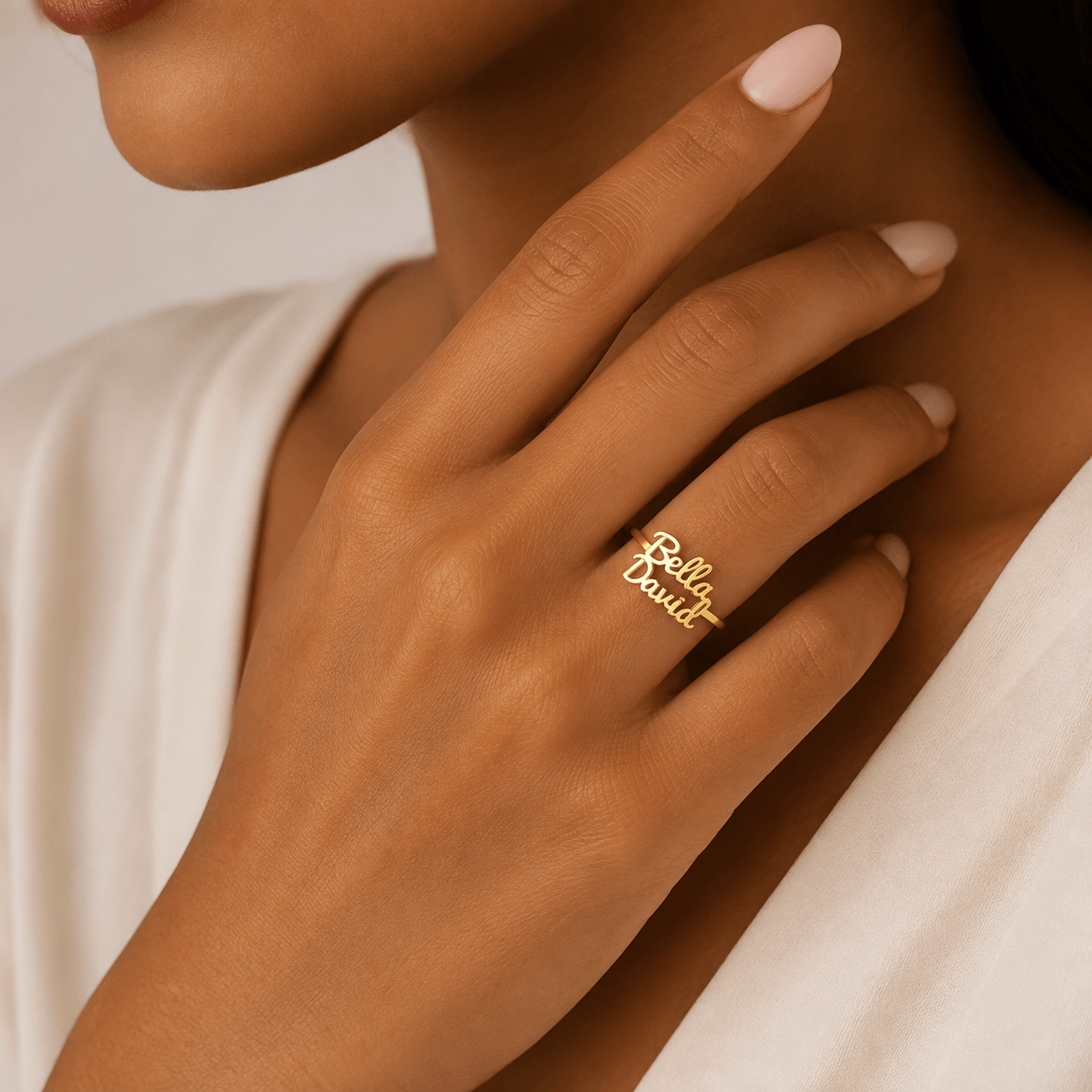A close-up lifestyle shot of a model's hand wearing a delicate gold script ring that reads 'Rex & Ruby'. The model has light pink manicured nails and is wearing a white top, set against a soft, warm neutral background. at shine gold and diamonds