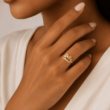 A close-up lifestyle shot of a model's hand wearing a delicate gold script ring that reads 'Rex & Ruby'. The model has light pink manicured nails and is wearing a white top, set against a soft, warm neutral background. at shine gold and diamonds